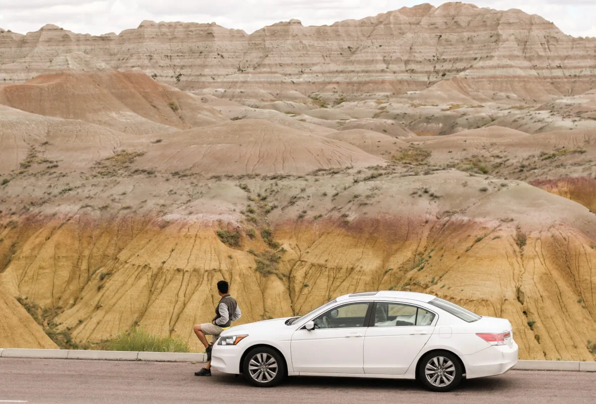Tyler La leaning against a white car at Badlands National Park, facing the layered rock formations during the 2018 road trip