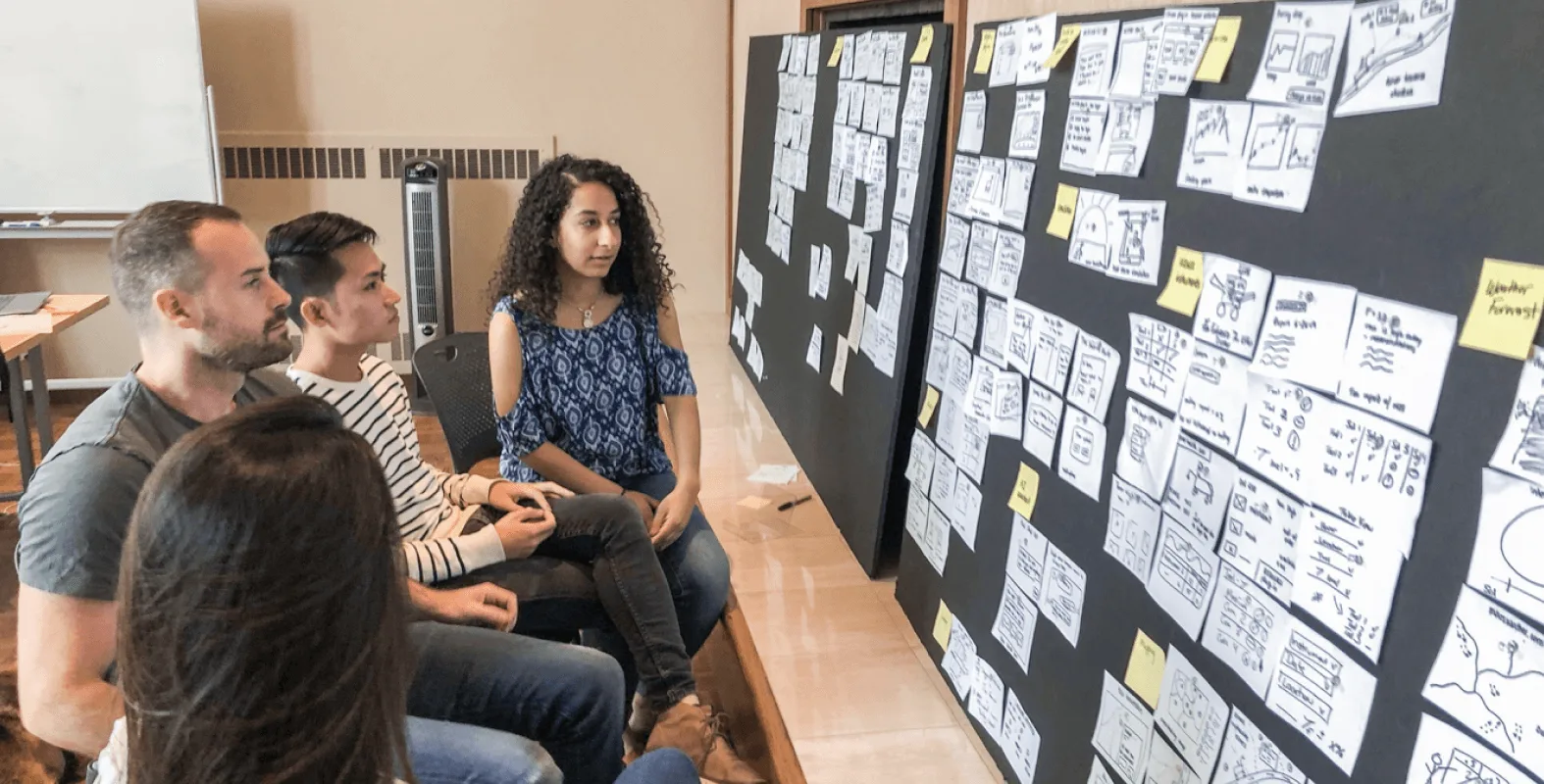 Four team members seated and reviewing a wall of hand-sketched wireframes and concepts pinned to black foam boards during an ideation session